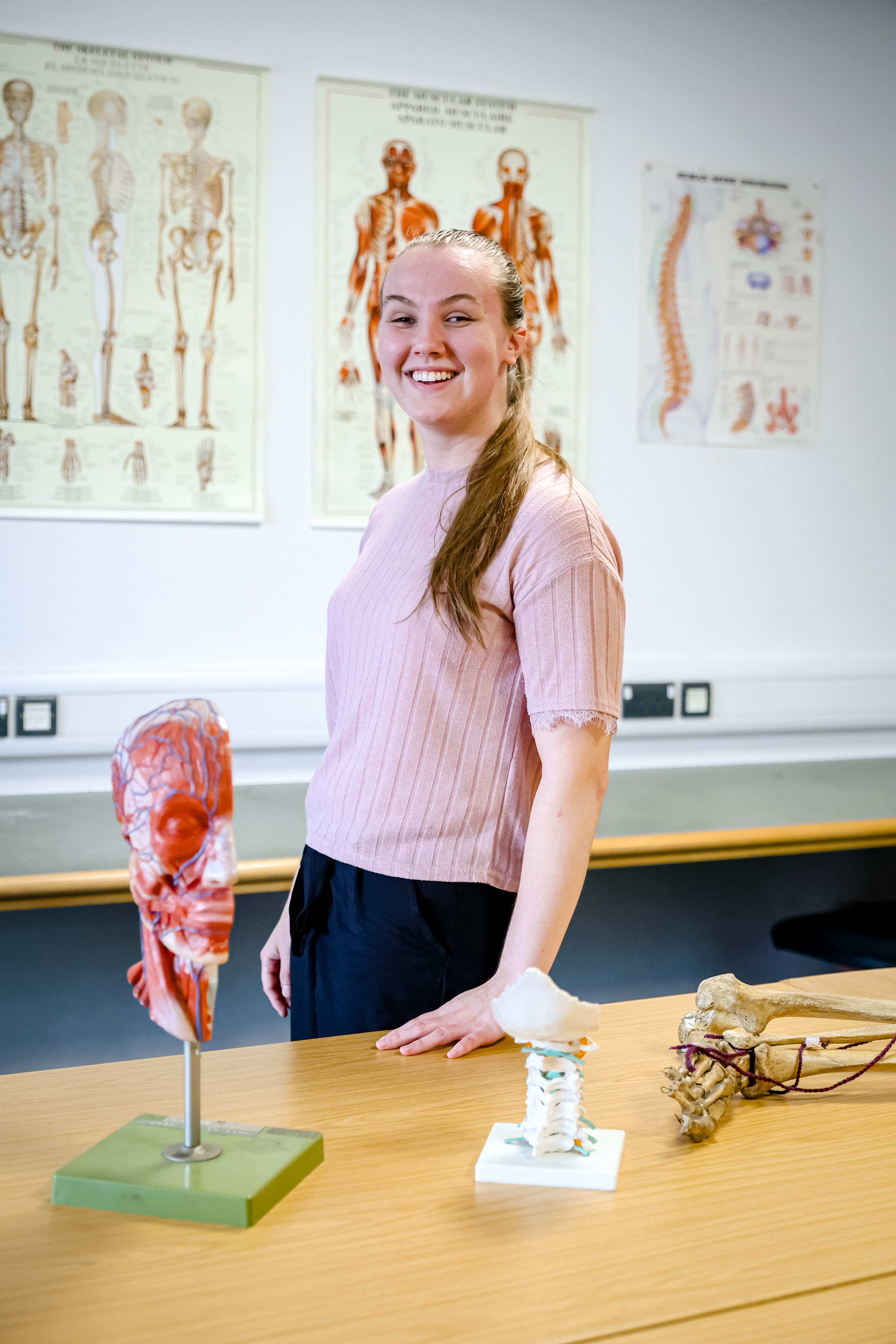 Lydia standing in the Massage classroom - there are models of the various sections of the body on the table in front of her and there are posters of the human body on the walls behind her 