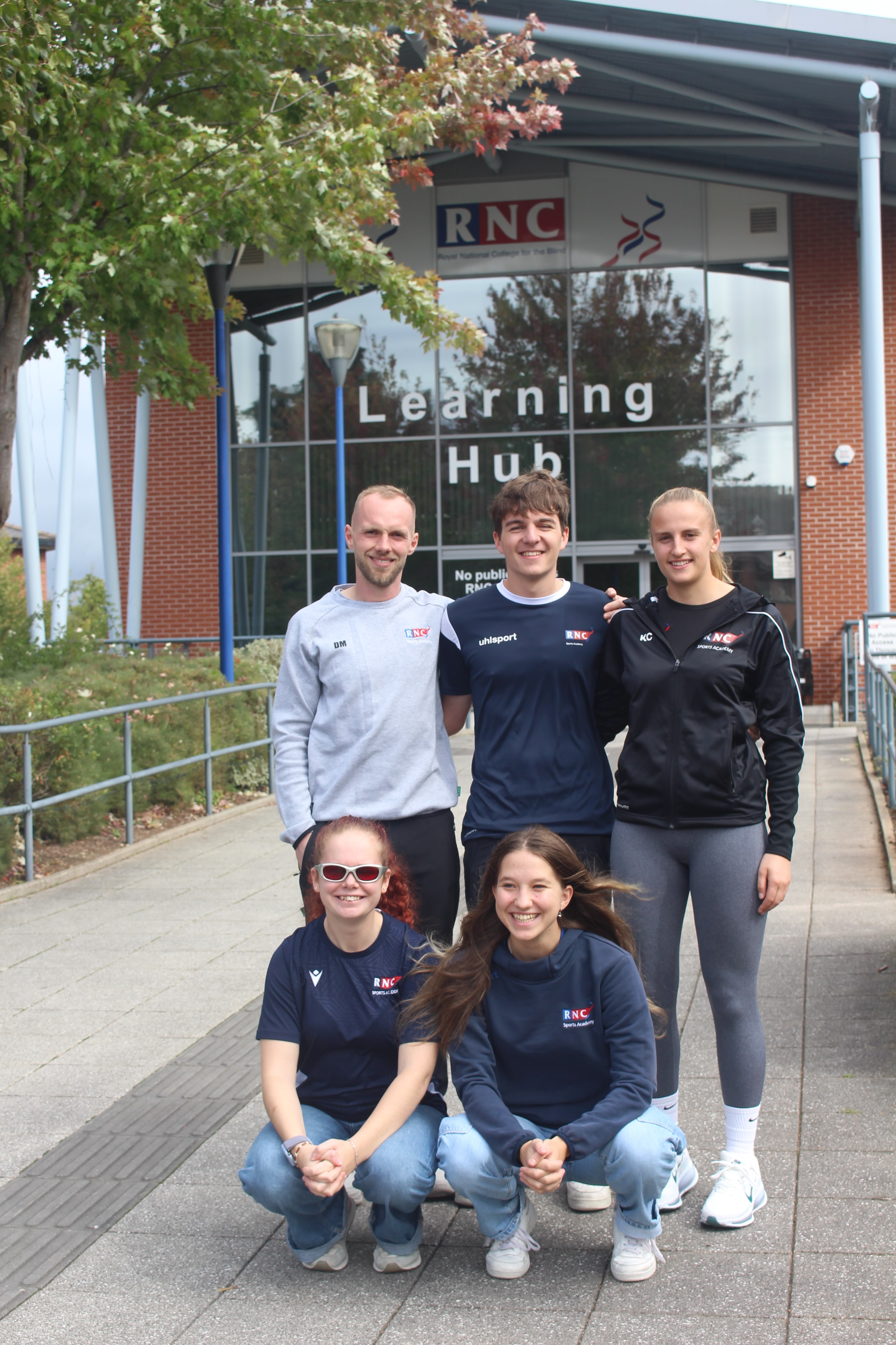 5 people assembled in front of the College's Learning Hub 3 stand and 2 crouch in front 