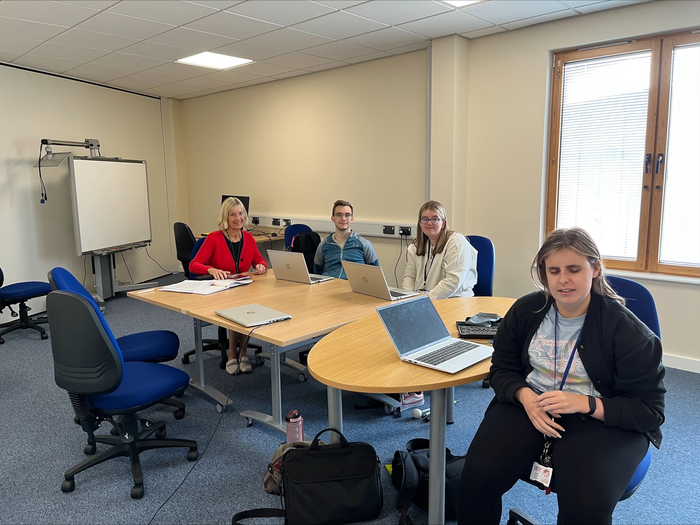 four people sitting around a table in a classroom setting. Three students have their laptops open in front of them on the table