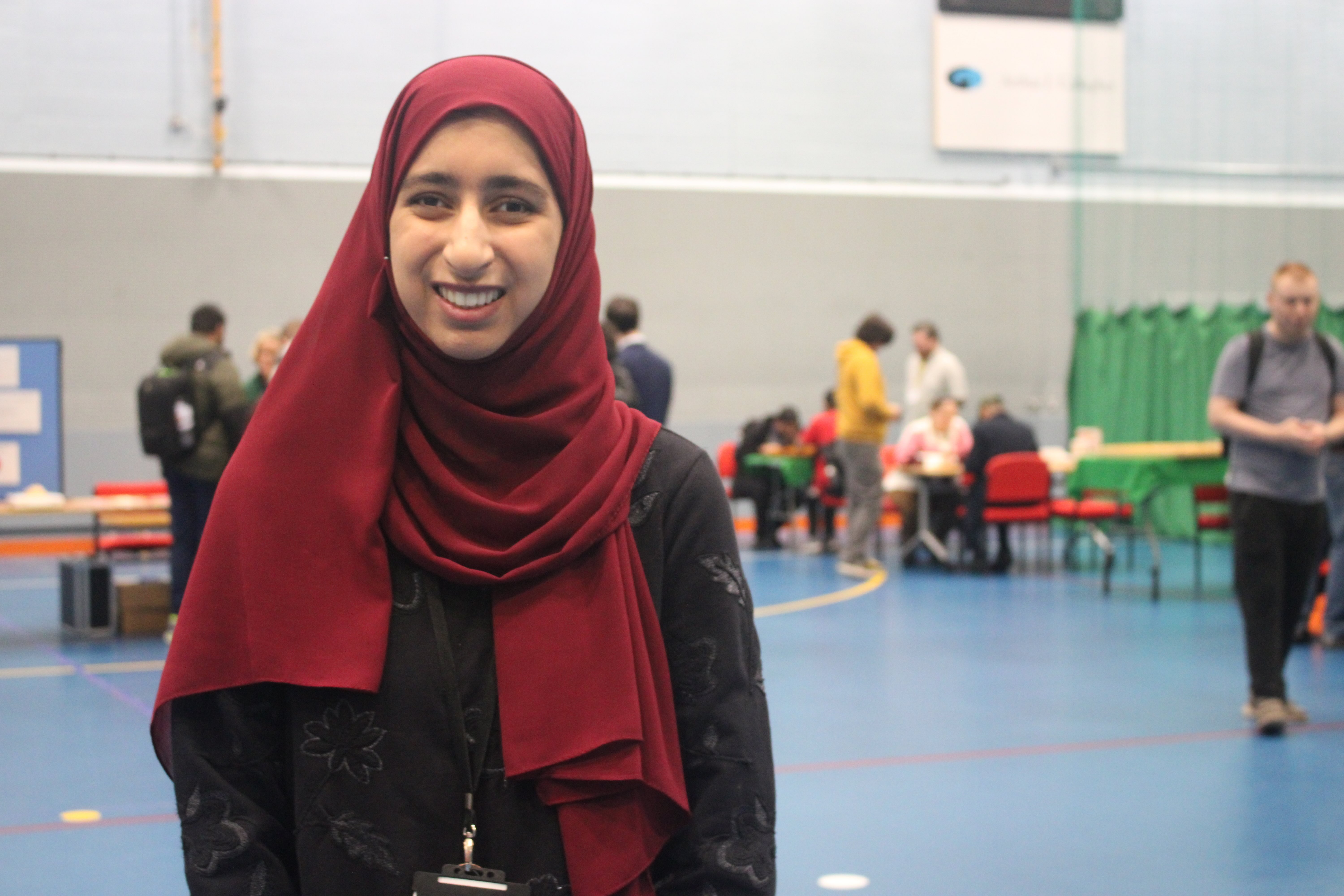 Humna wearing a burgundy coloured headscarf and a black sweatshirt is pictured in the centre of the hall with stands and visitors milling around her