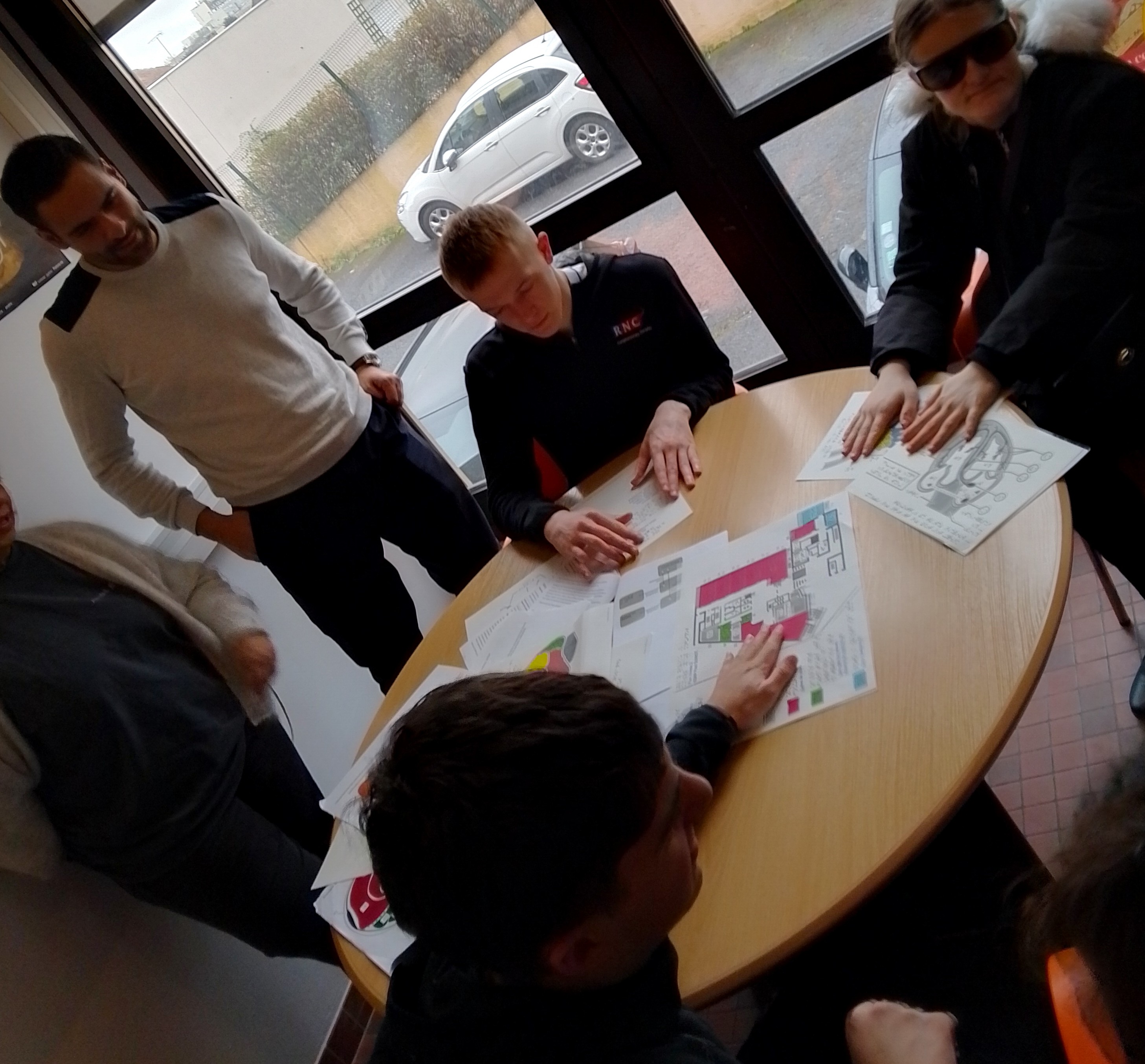 A group of students seated around a table exploring tactile diagrams of the anatomy a member of the French staff stands over them 
