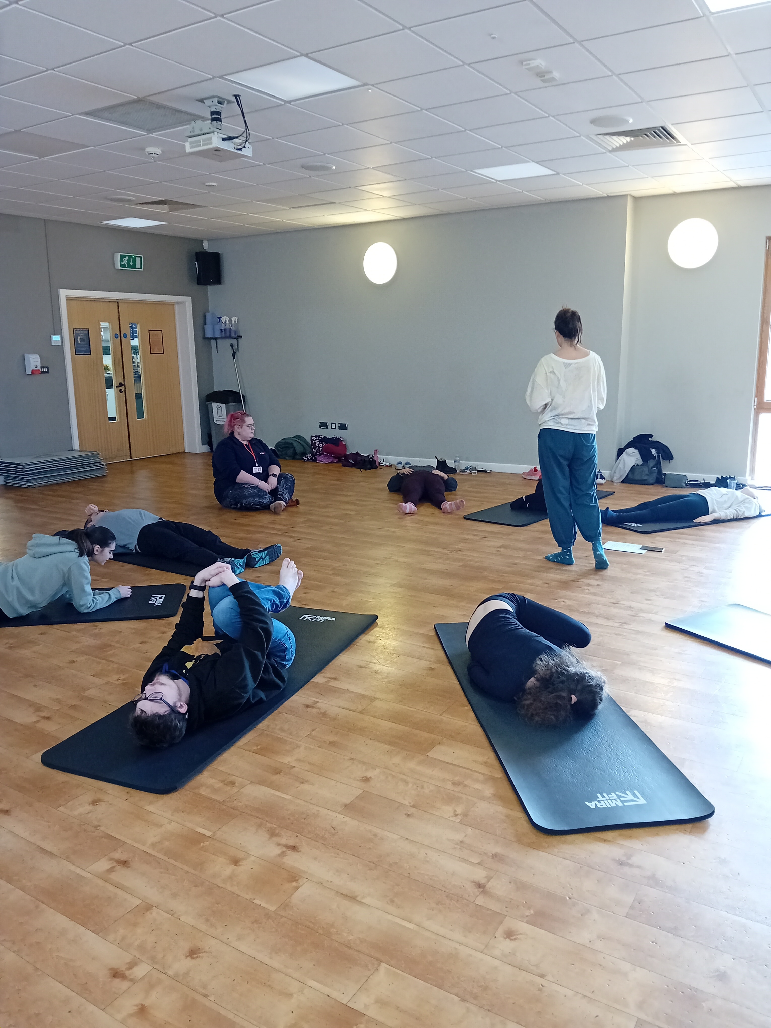 Students lie on mats in a circle, they are all adopting very different poses as the teacher stands guiding them