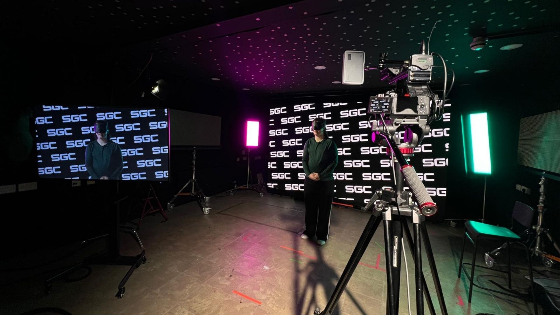 Leah stands in a dark studio wearing Goalball shades he is surrounded by large screens with brightly coloured graphics and cameras 