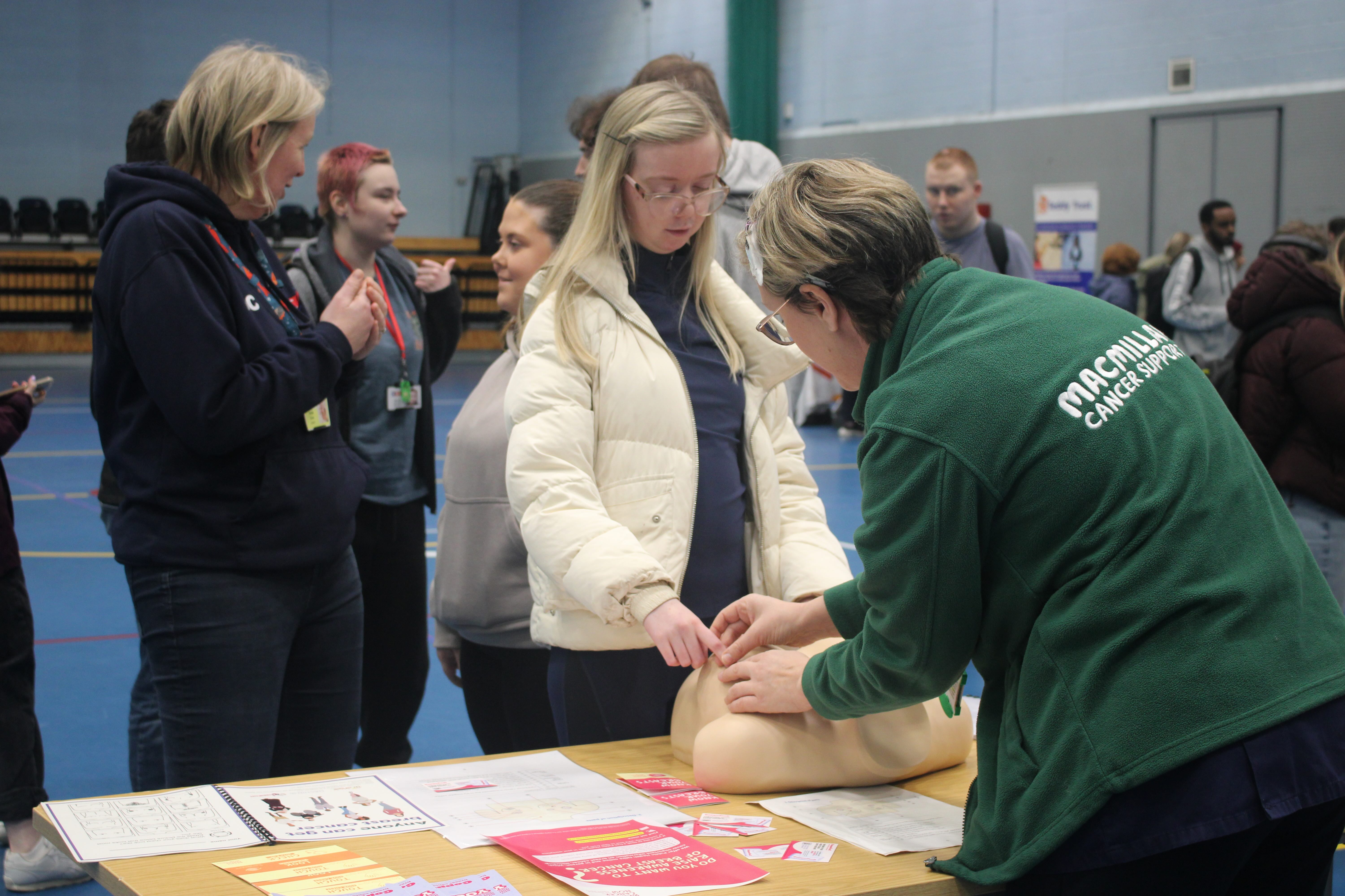 the Macmillan Nurse guides a female students fingers over a pair of prosthetic breasts to help identfiy the signs of breast cancer