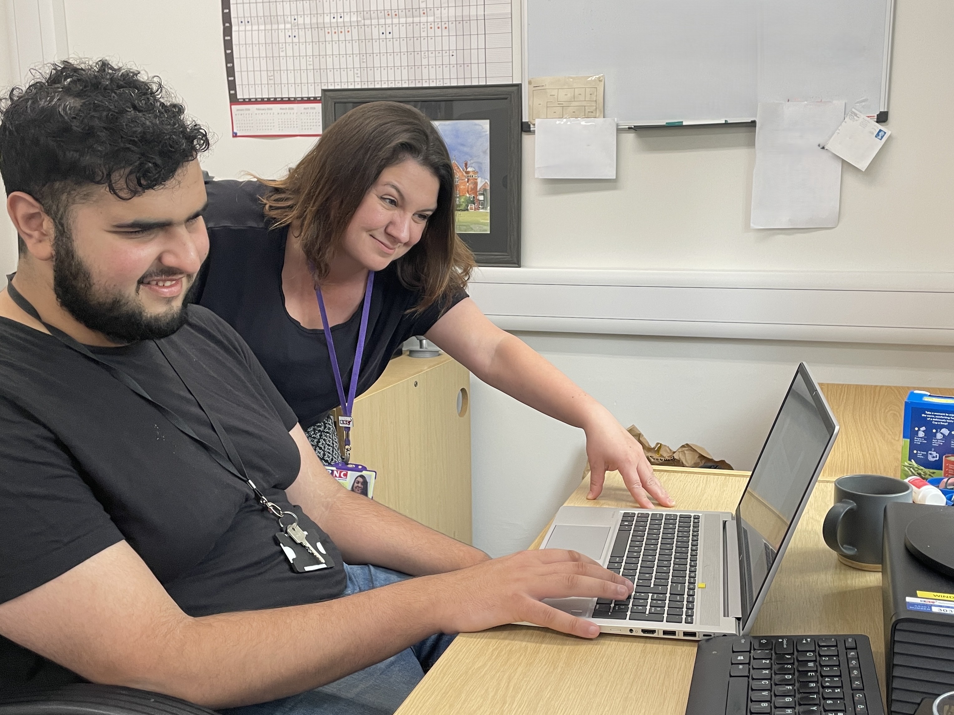 two people at a desk one is working on a laptop as the other leans over appearing to be giving advice