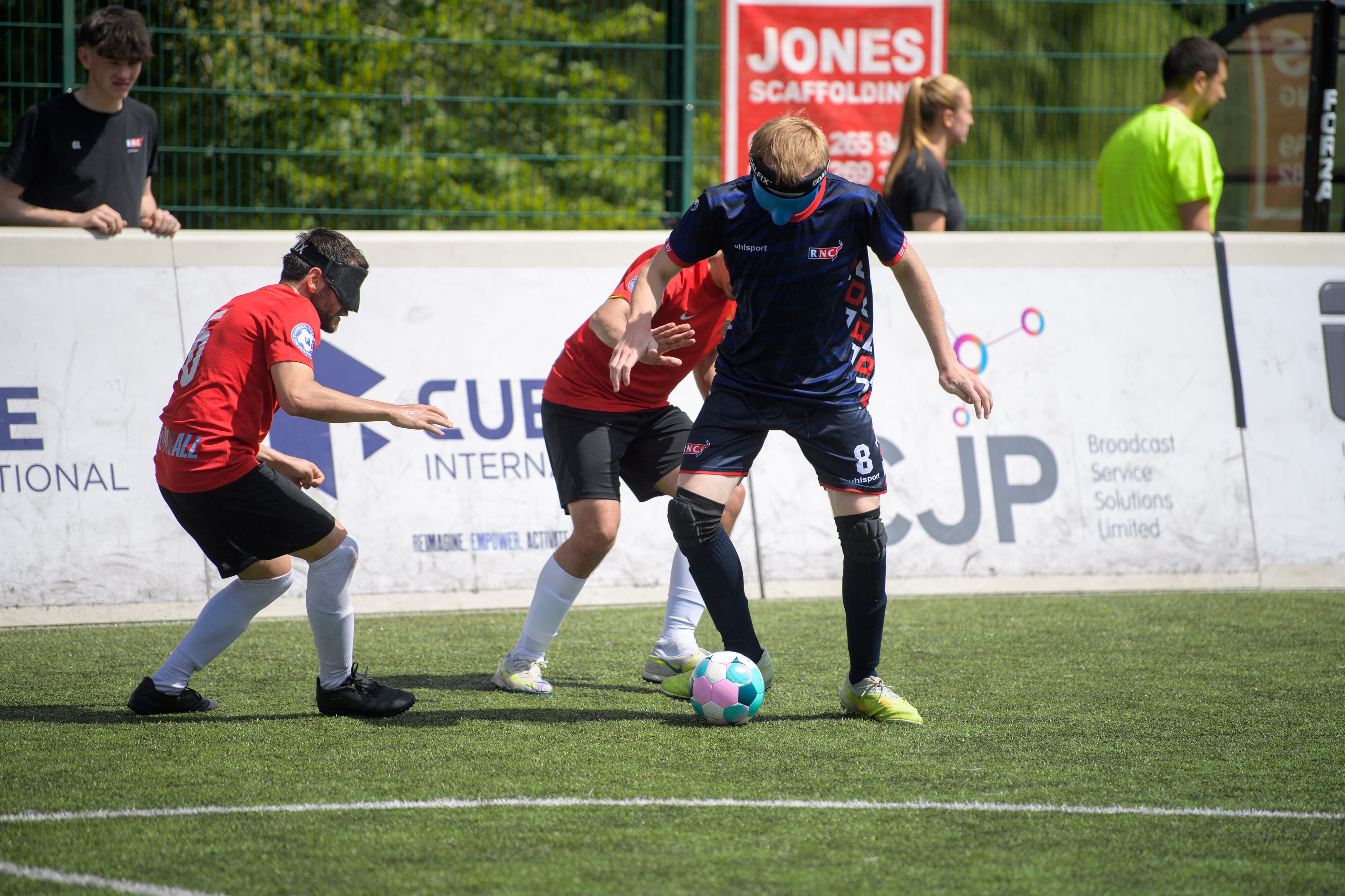 An RNC player on the ball wearing RNC's navy blue strip. There are two Pirsos players around him wearng a red strip 